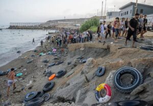 Толпа мигрантов на захламлённом пляже с шинами и спасательным кругом у скалы